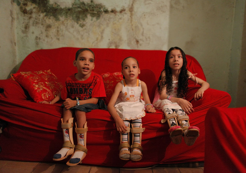 The children of the Amor Divino family, Dhones, Izabely and Samille, sit on their couch in Sao Paulo