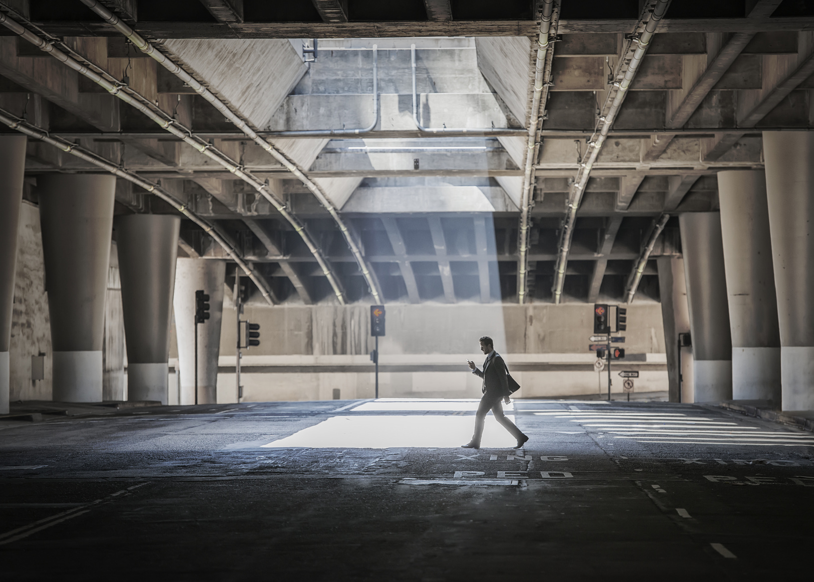 A person crossing an open space, a city underpass, with concrete walls and a pool of sunlight.